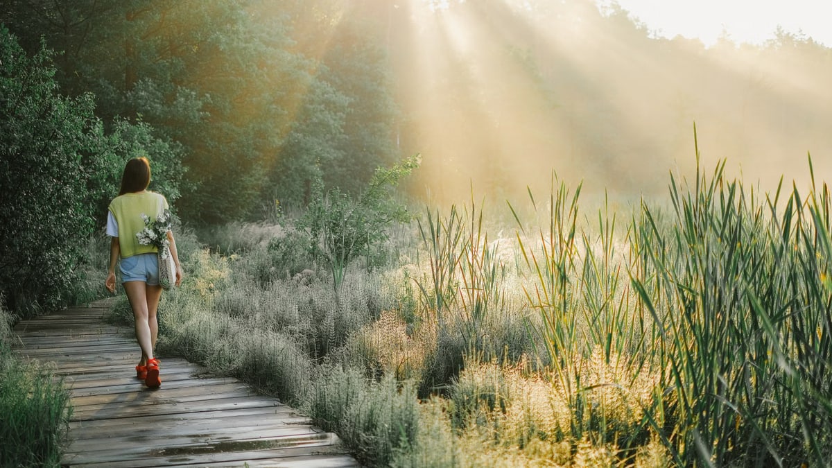 A woman enjoys a peaceful walk on a wooden path in a sunlit forest, surrounded by lush greenery.